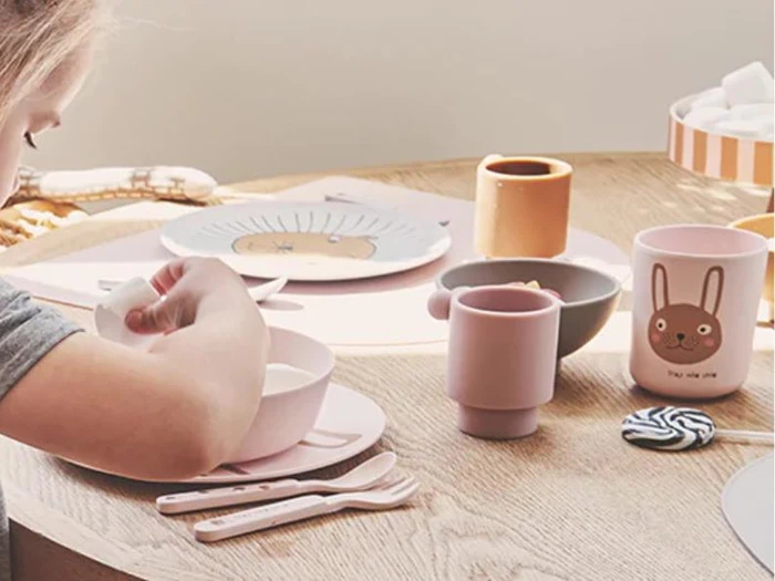Et barn sidder ved et træbord med pastelfarvede tallerkener med dyreansigter, kopper, skåle og redskaber, der er arrangeret pænt til et måltid.