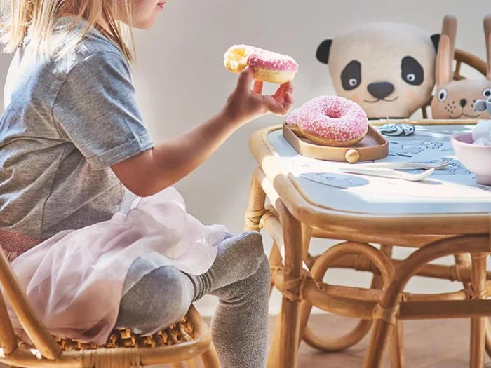 Et lille barn sidder ved et lille bord med en lyserød donut i hånden, en anden donut på en tallerken og udstoppede panda- og hundelegetøj på bordet.