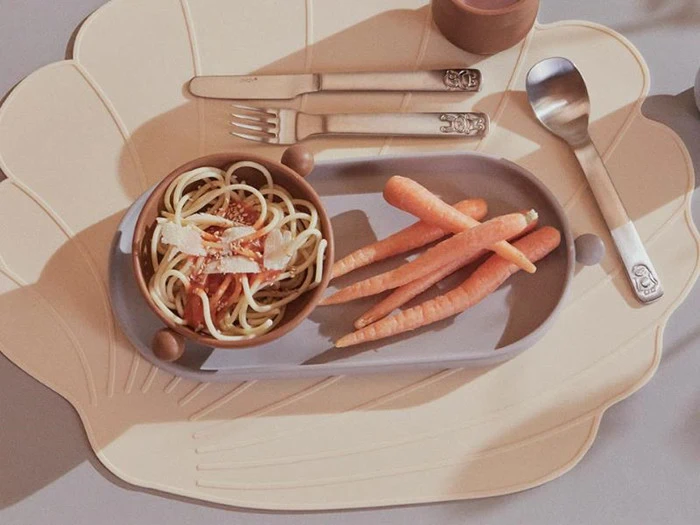 En tallerken med flere babygulerødder og en lille skål spaghetti med ost, sat på en skalformet dækkeserviet sammen med en gaffel, en ske og en kniv.