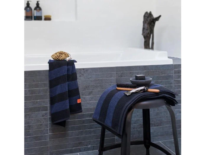 Et moderne badeværelse med et gråt flisebadekar, et stribet blåt og sort håndklæde på kanten og en skammel med lignende håndklæder, en børste, en barbermaskine og en sæbeskål.