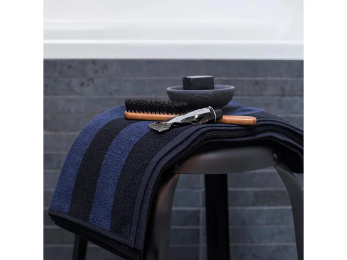 Et foldet marineblåt og sortstribet håndklæde på en skammel med en barberkost, sæbe og barberkniv placeret ovenpå. Mørk flisebelagt væg og badekar i baggrunden.
