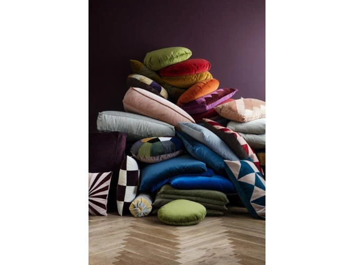 En bunke farverige, blandede hynder og puder stablet op mod en mørklilla væg på et sildebensgulv af træ.