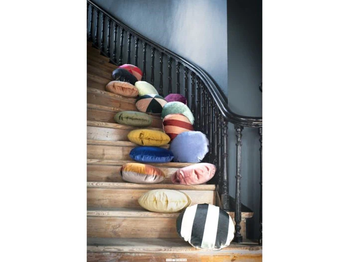 En samling farverige runde puder er spredt langs en trætrappe med et mørkt gelænder i et hus med grå vægge.