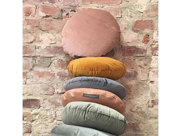 Seks runde meditationspuder i forskellige farver er stablet lodret op ad en rustik, eksponeret murstensvæg.