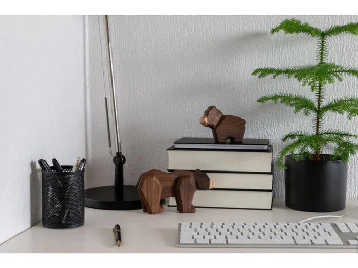 Et minimalistisk skrivebord med bøger, en lampe, en potteplante, kuglepenne i en holder, bjørnefigurer i træ, en kuglepen og et hvidt tastatur.