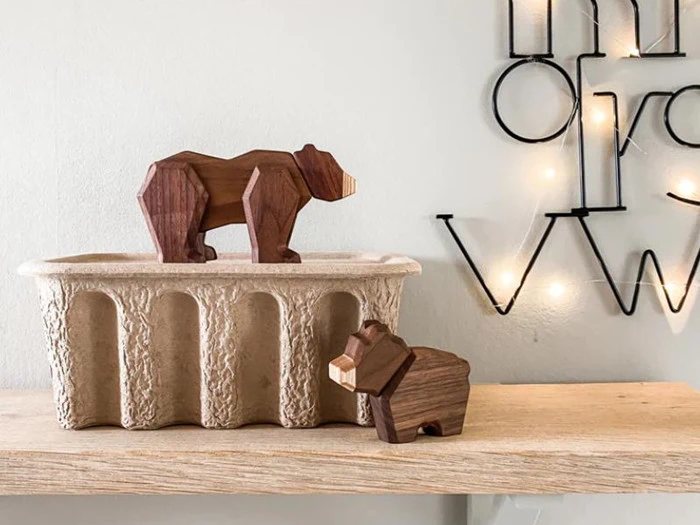 To geometriske bjørnefigurer af træ på en træhylde; den ene står på en tekstureret beholder, den anden ved siden af. I baggrunden ses et dekorativt vægstykke med lyskæder.