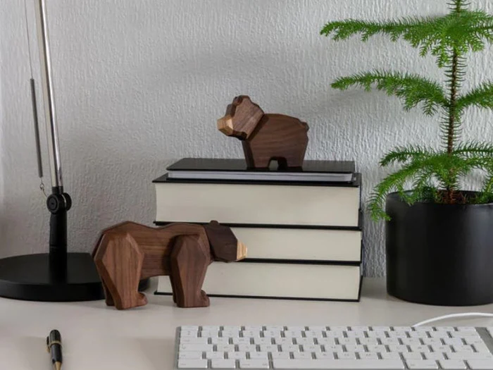 Et skrivebord med et tastatur, stablede bøger, to bjørnefigurer i træ, en sort lampe og en potteplante mod en lys væg.