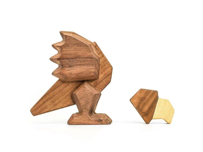 To minimalistiske, geometriske træfigurer på en hvid baggrund; den ene ligner en fugl, den anden et lille dyr, muligvis en kylling, og begge er udformet i naturlige træfarver.