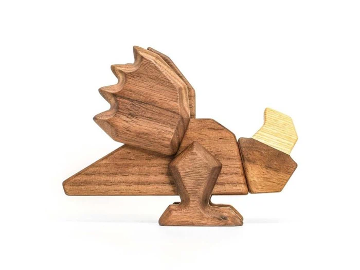 Et stykke trælegetøj formet som en drage med kantede vinger, ben og hale, lavet af forskellige nuancer af træ på en hvid baggrund.