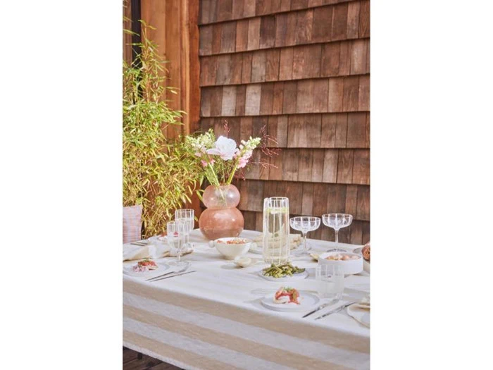 Et bord dækket til et måltid med glas, tallerkener med mad og en lyserød vase med hvide blomster, udendørs ved siden af en væg af træspån og grønne planter.