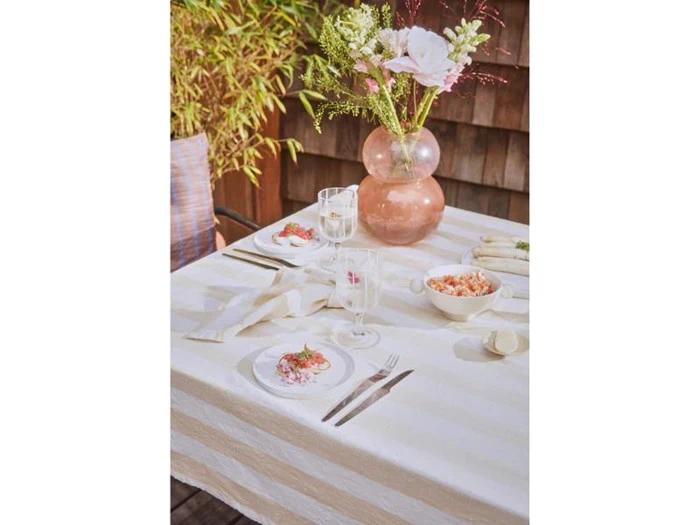 Et bord dækket til to med hvide tallerkener, bestik, glas, en blomsterdekoration i en lyserød vase og skåle med mad på en hvid dug, udendørs med planter i baggrunden.