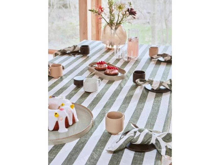 Et bord dækket med en grøn- og hvidstribet dug, desserter, krus, servietter og en vase med blomster ved et vindue med udsigt til naturen.