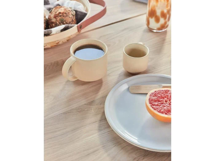 Et beige kaffekrus, en lille kop, en halv grapefrugt på en tallerken med en træspatel og en kurv med brød på et lyst træbord.