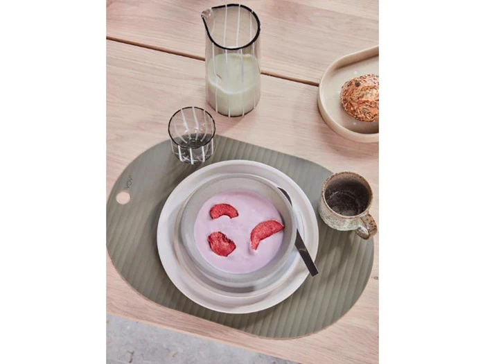 En skål lyserød yoghurt med jordbær i skiver, en ske, en kop, et glas vand, en kande mælk og et rundstykke på et træbord med dækkeserviet.