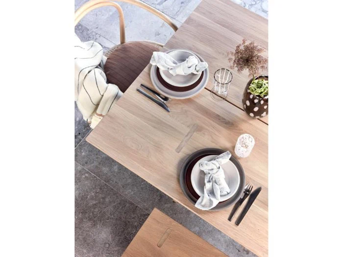 Et spisebord i træ til to personer med tallerkener, bestik og servietter, en stol med et stribet tæppe og små vaser med blomster.