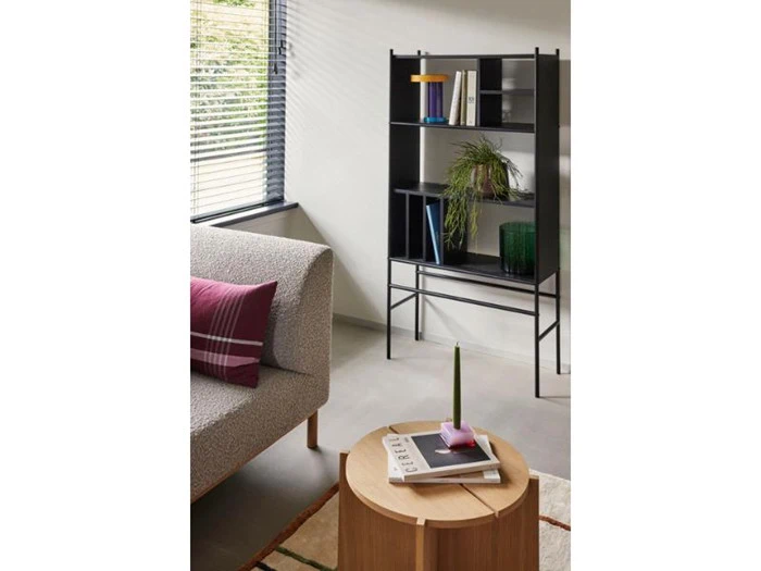 En moderne stue med en grå sofa, et rundt sofabord i træ og en sort metalreol med bøger, dekorationer og en potteplante nær et vindue med persienner.