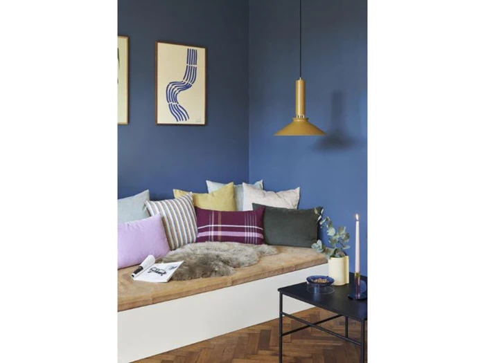 En hyggelig krog med en træbænk, forskellige farverige puder, en pelskvast, blå vægge, moderne kunst, en gul pendel og et lille sort bord med dekoration.