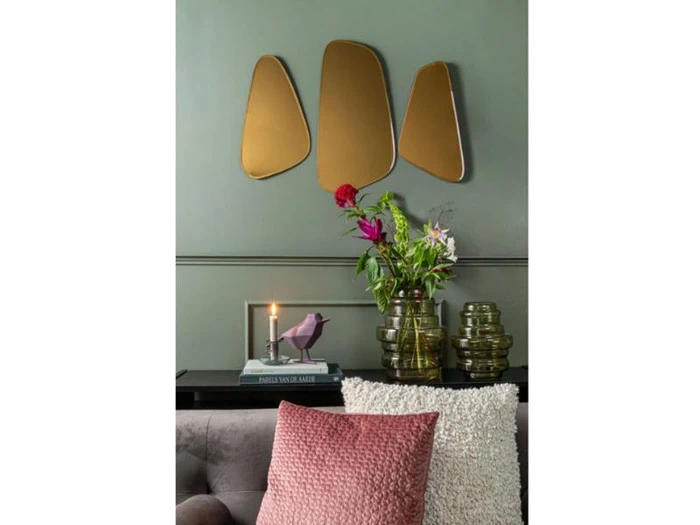 En moderne stue med grønne vægge, abstrakte guldspejle, en vase med blomster, stablede bøger, en fuglefigur og strukturerede rosa og cremefarvede puder på en grå sofa.