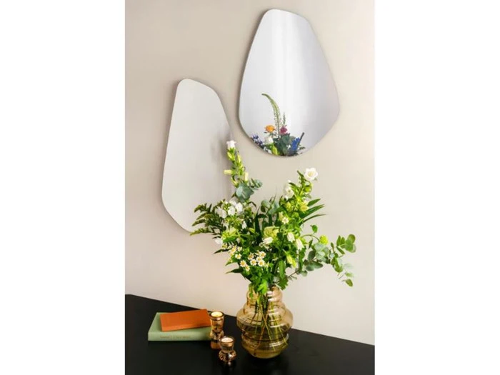 En glasvase med grønne og hvide blomster står på et sort bord med to uregelmæssigt formede spejle og et par bøger og små krukker ved siden af. Blomsterne spejler sig i spejlene.