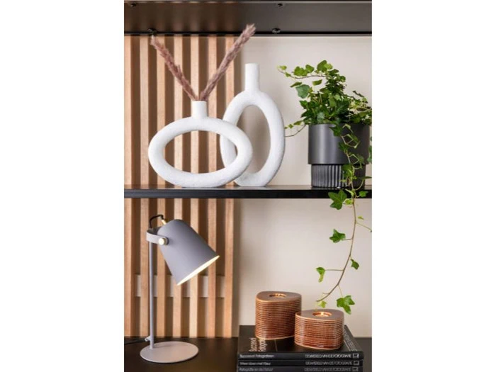 En moderne hylde viser to hvide abstrakte vaser, en vedbendplante i potte, en grå skrivebordslampe, to trælysestager og stablede bøger mod en baggrund af trælameller.