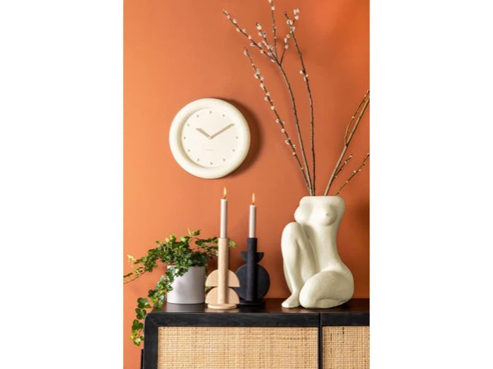 En moderne indretningsscene med et rundt vægur, stearinlys, en plante og en abstrakt hvid skulptur på et skab mod en orange væg.