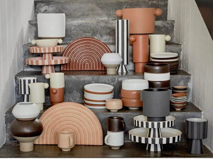 Farverige, geometriske keramikvaser, -tallerkener og -krus er arrangeret på betontrapper i en moderne, kunstnerisk udstilling. Mange af genstandene har striber og jordfarver som hvid, sort, brun og te