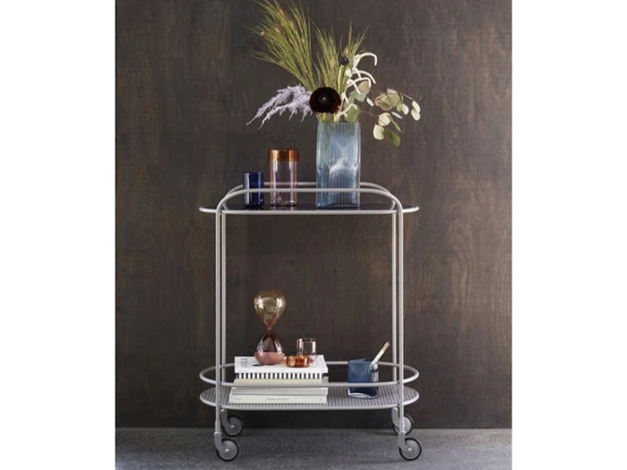 En sølvfarvet barvogn med to hylder rummer vaser med planter, glas, bøger og pyntegenstande mod en mørk væg med tekstur.