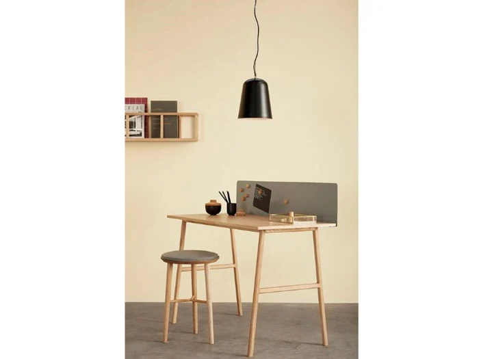 Minimalistisk arbejdsplads med skrivebord og skammel i træ, en lille hylde med bøger, en skillevæg med noter og en sort pendellampe over.