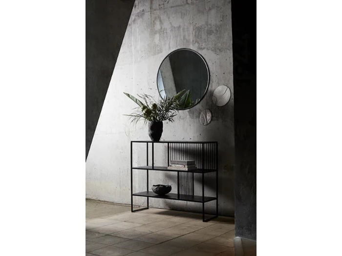 En moderne entré med et rundt spejl, en betonvæg, et konsolbord i metal, en potteplante og en dekorativ skål, oplyst af naturligt lys.