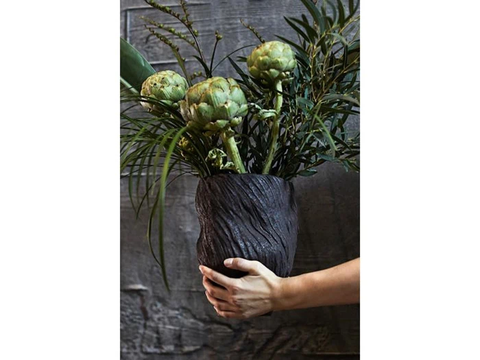 En hånd holder en mørk, struktureret vase fyldt med artiskokker og grønne blade foran en rustik trævæg.
