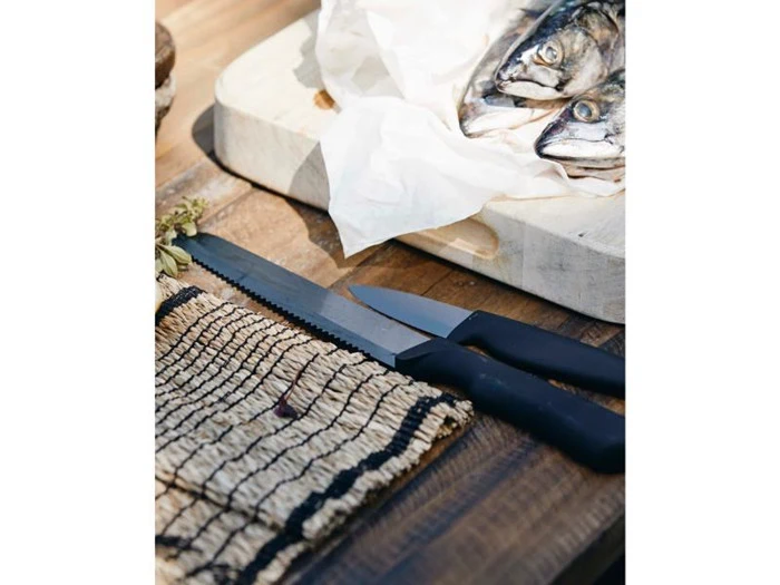 To knive med sorte skafter ligger på et træbord ved siden af en vævet måtte, tæt på et skærebræt med to fisk pakket ind i hvidt papir.