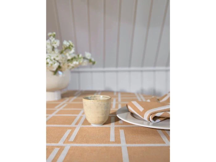 En beige kop står på en mønstret dug ved siden af en foldet serviet på en hvid tallerken med en hvid blomstervase i baggrunden.