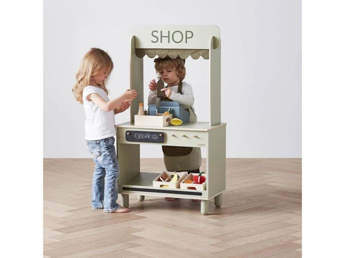 To små børn leger med en legetøjsbutik og lader, som om de køber og sælger madvarer af træ i et rum med lyst trægulv.