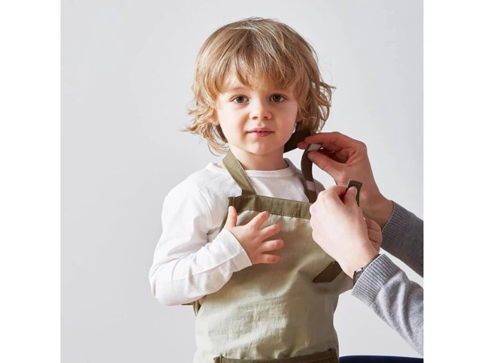 Et lille barn iført et khakifarvet forklæde får justeret forklædestroppen af en voksen på en almindelig lys baggrund.