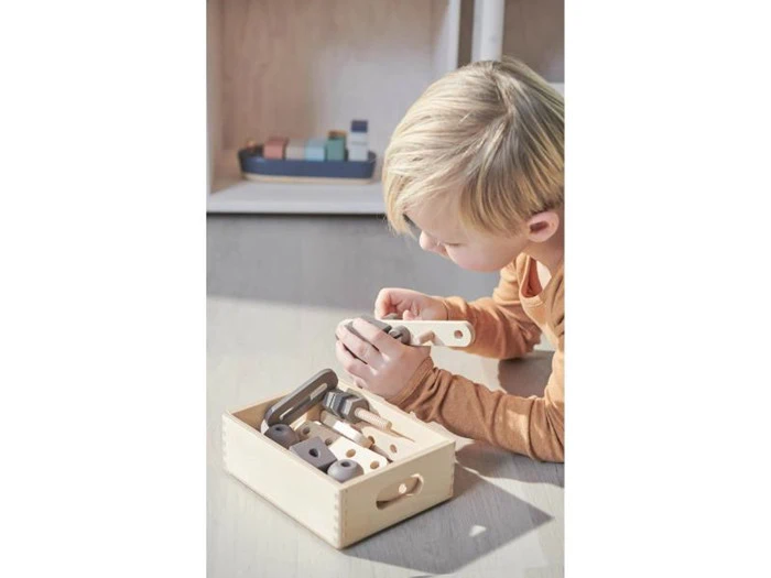 Et lille barn med lyst hår ligger på gulvet og leger med legetøjsredskaber af træ fra en lys kasse.