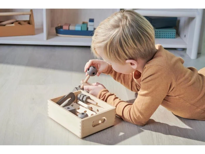 Et lille barn i brun pyjamas ligger på et trægulv og leger med et værktøjssæt af træ fra en kasse, mens det samler delene med fokuseret opmærksomhed.