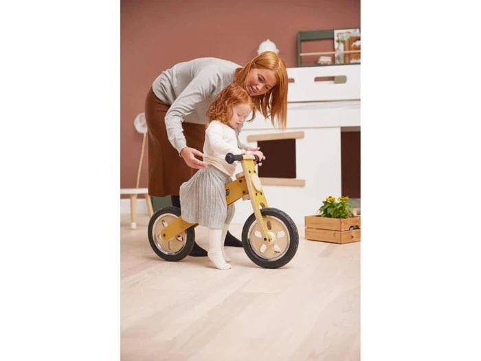 En voksen hjælper et lille barn med at køre på en gul løbecykel indendørs. Barnet har en lys kjole og trøje på, og scenen er blødt og varmt belyst.