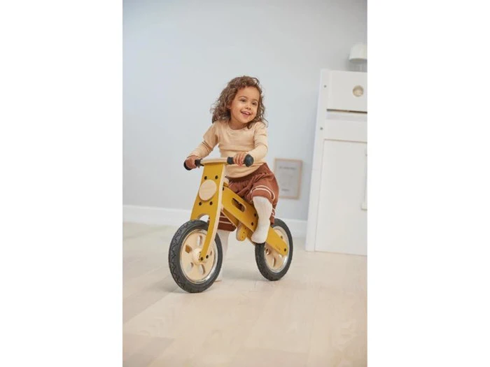 Et lille barn med krøllet hår smiler, mens det kører på en gul løbecykel indendørs på et lyst trægulv.