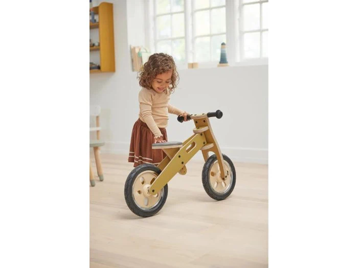 Et lille barn smiler, mens det står ved siden af en løbecykel af træ i et lyst, solbeskinnet rum med lyse gulve.