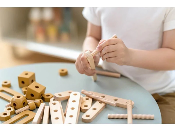 Et barn i hvid skjorte samler byggelegetøj af træ med forskellige stænger, klodser og plader på et lyseblåt bord.