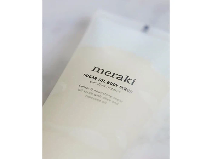 Nærbillede af en Meraki Sugar Oil Body Scrub-tube med tekst, der beskriver den som en certificeret økologisk, skånsom og nærende sukkeroliescrub.