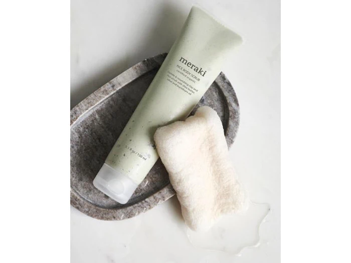 En tube Meraki facial exfoliator ligger på en stenbakke ved siden af en hvid, struktureret svamp, alt sammen på en hvid overflade.