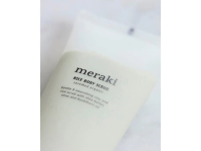 Nærbillede af en hvid tube mærket meraki Rice Body Scrub, der beskrives som en blid og nærende certificeret økologisk scrub med hvidt ler og naturlige olier.
