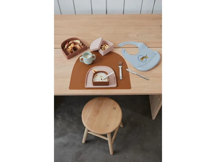 Et træbord med en skammel, børnemad i opdelte tallerkener, et krus, en gaffel, en blå hagesmæk og snacks i små beholdere.