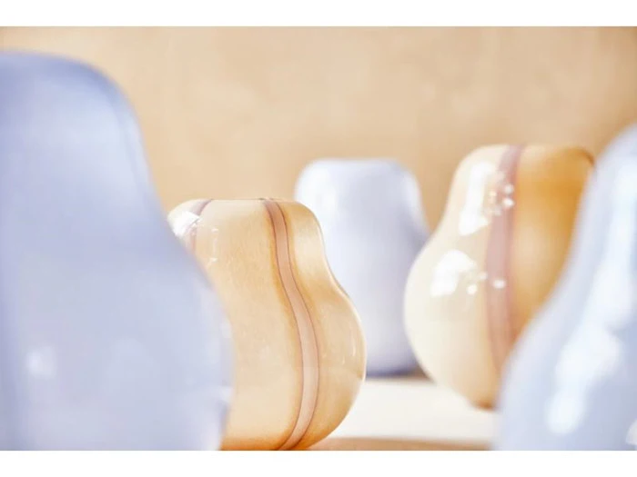 Flere glatte, abstrakte glasvaser i bløde pastelfarver af blå og beige er arrangeret på en lys overflade med en sløret baggrund.