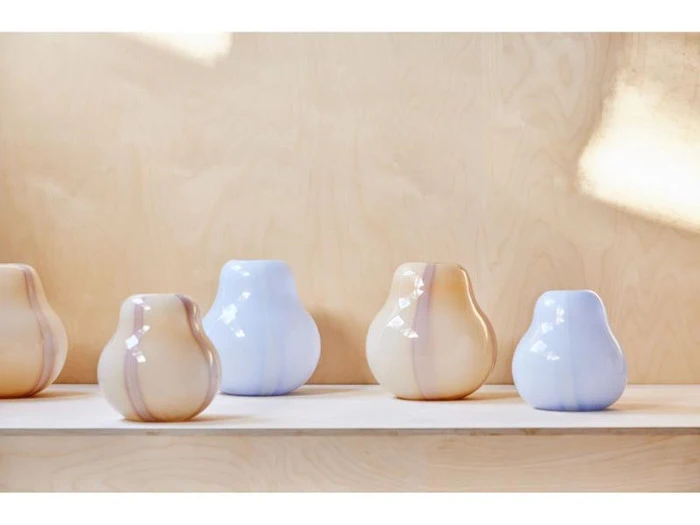 Fem glatte, afrundede keramikvaser i pastelblå og beige står på en lys træhylde mod en beige væg.