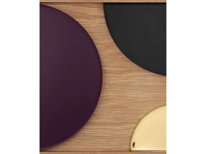 Tre geometriske former - en lilla halvcirkel, en sort halvcirkel og en kvartcirkel i guld - er placeret på en lys træoverflade.