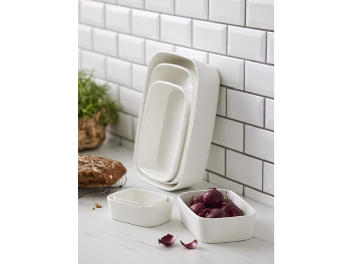 Fire hvide keramiske bageforme i forskellige størrelser er stablet og arrangeret på et hvidt køkkenbord ved siden af brød, løg og en potteplante med en hvid flisebelægning bagved.