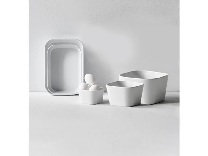 Et sæt minimalistisk hvidt keramisk køkkenudstyr, herunder en bakke, to skåle og en kop med tre æg, arrangeret på en hvid overflade mod en hvid baggrund.