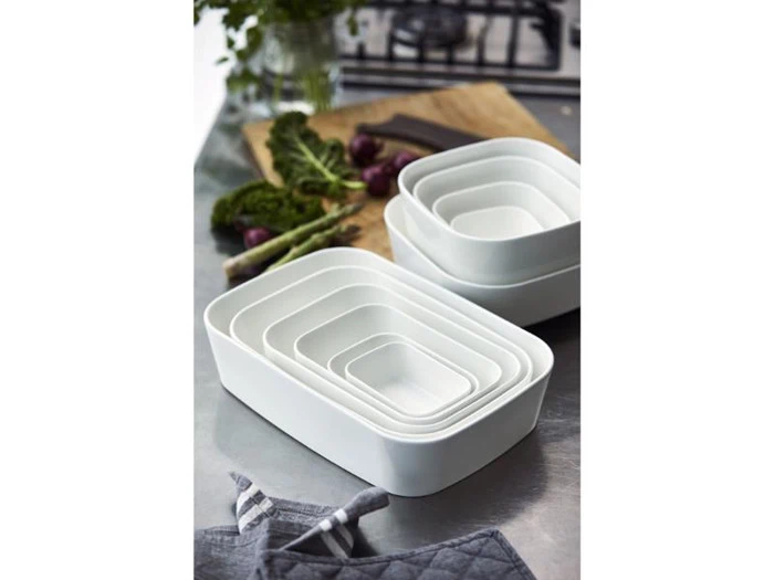 Et sæt af seks hvide rektangulære redefade på et køkkenbord med grøntsager og et skærebræt i baggrunden.